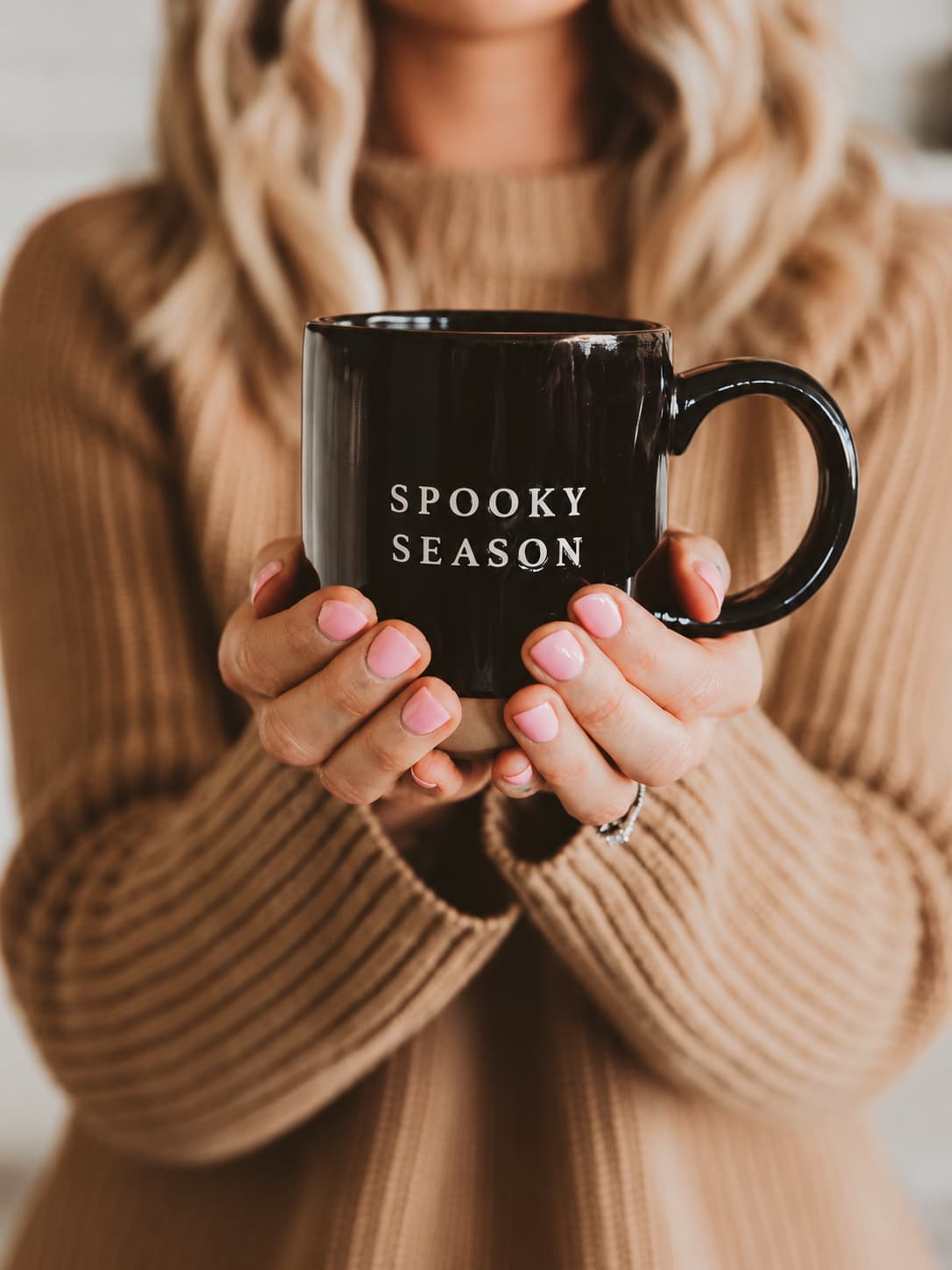 Woman wearing tan sweater holding black stoneware mug with the words "spooky season" on it.
