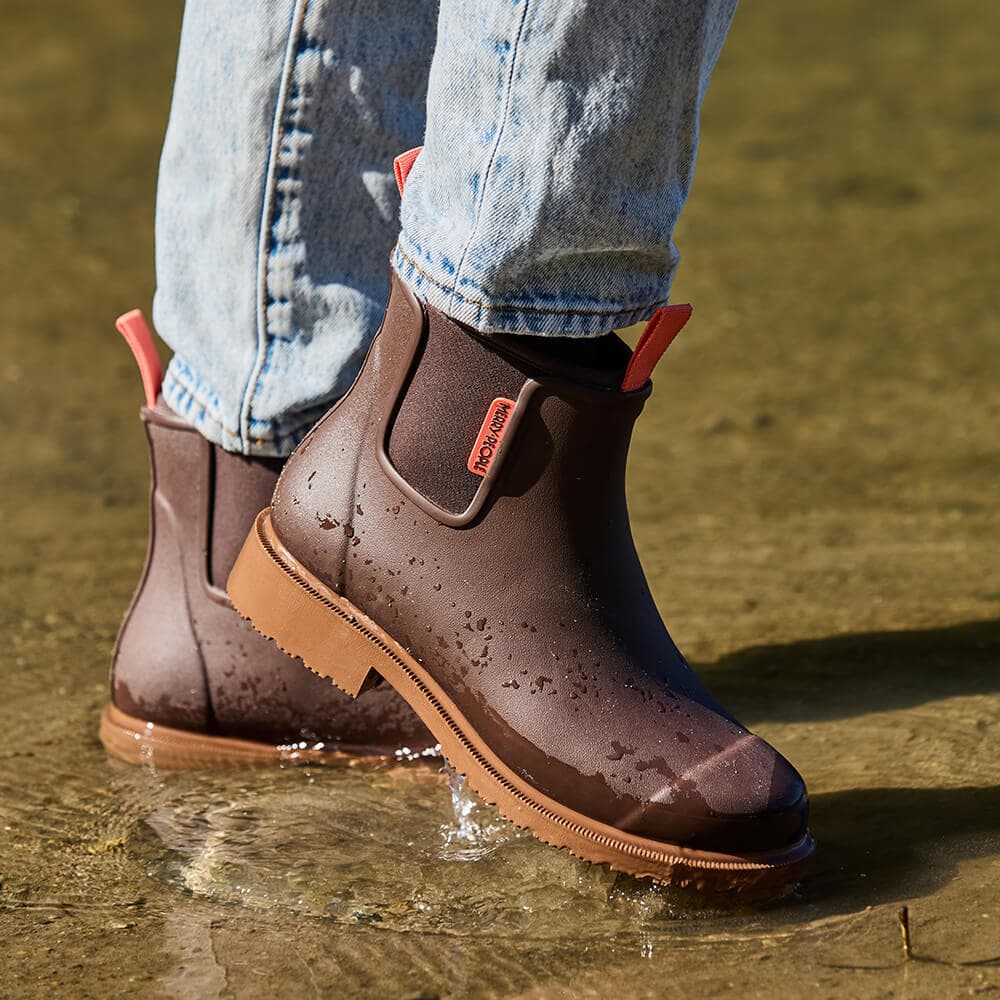 A person wearing dark brown waterproof Chelsea-style rain boots with elastic side panels and pull tabs, standing in shallow water with light blue jeans visible.