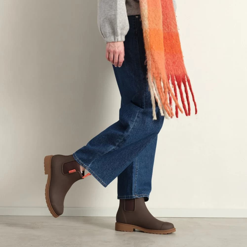 Person wearing brown boots, blue jeans, and an orange scarf on a neutral background