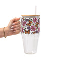A hand holding a clear glass tumbler with a bamboo lid and straw. The tumbler features a colorful wrap-around pattern of orange monarch butterflies, pink and purple flowers, and green leaves on a white background.