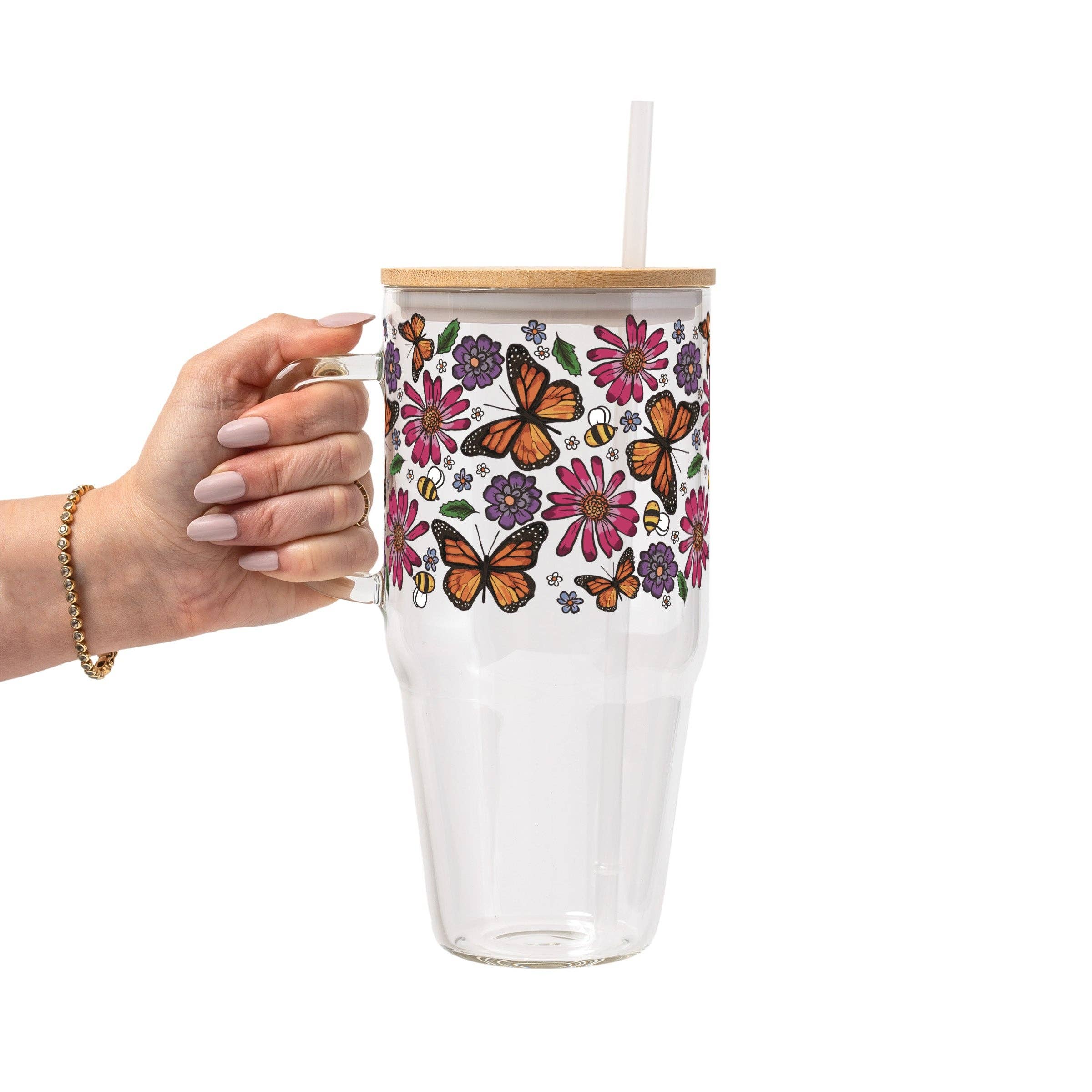A hand holding a clear glass tumbler with a bamboo lid and straw. The tumbler features a colorful wrap-around pattern of orange monarch butterflies, pink and purple flowers, and green leaves on a white background.