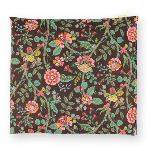 Small patterned hot or cold aromatherapy wrap with botanical pattern with pink, yellow, and green flowers and leaves on a dark brown background. A small cream-colored fabric tag is visible on the top right edge.