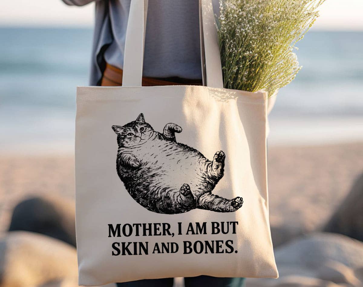 A cream canvas tote bag with a black graphic of a belly-fat cat and the text 'MOTHER, I AM BUT SKIN AND BONES.' being carried on a beach by a person.