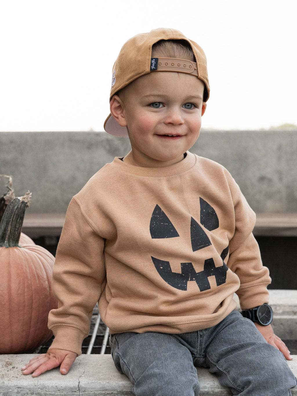 A young child wearing a tan crewneck sweatshirt with a black jack-o'-lantern face graphic, gray jeans, and a tan baseball cap, sitting outdoors next to a pumpkin.