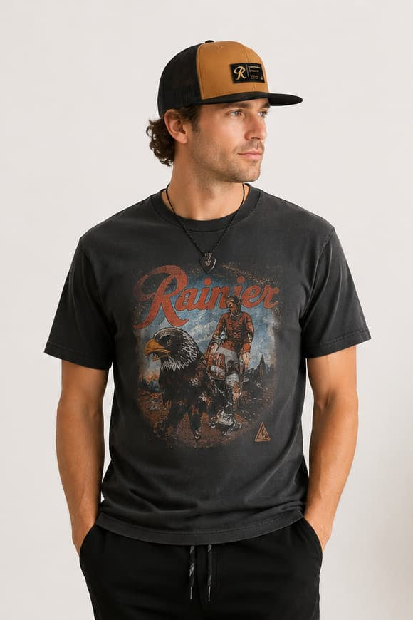 A man wearing a dark charcoal graphic t-shirt, black sweatpants, black and white sneakers, and a brown and black baseball cap, standing in a minimalist room with a potted plant.