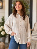 A woman wearing a cream and brown vertically striped button-down blouse with balloon sleeves, paired with blue jeans, standing on a porch.