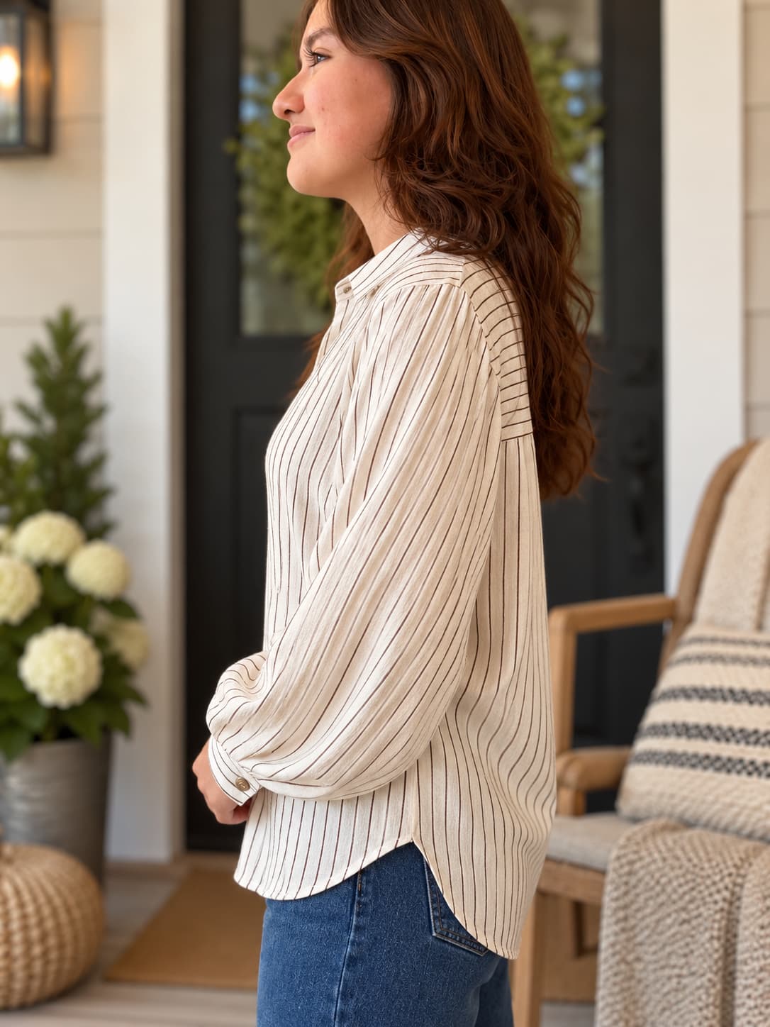 A woman wearing a cream-colored long-sleeve button-up blouse with thin vertical stripes, paired with blue jeans, standing on a porch.