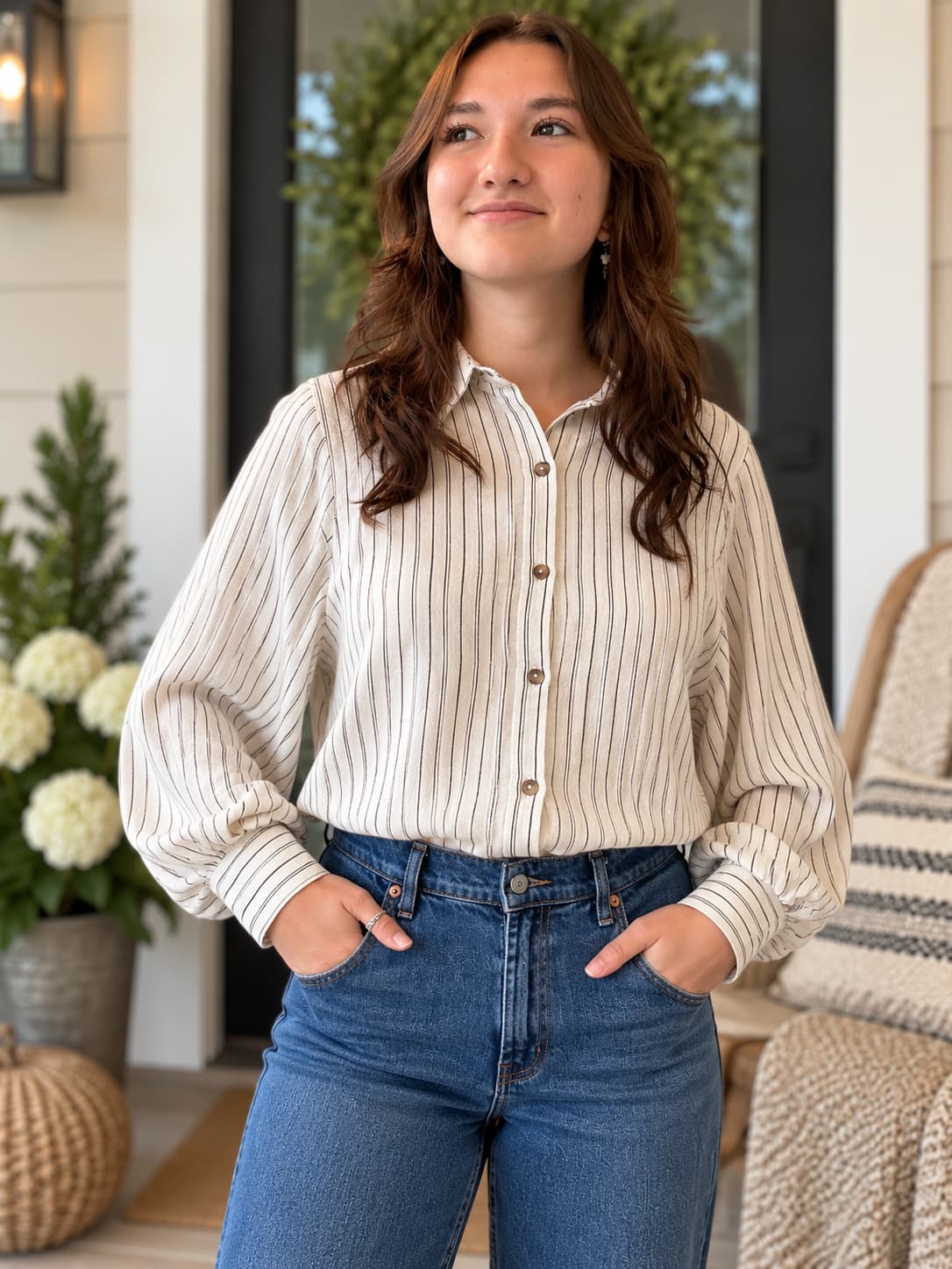 A woman wearing a cream-colored, long-sleeve button-up shirt with thin vertical brown stripes, paired with blue jeans, standing on a porch.