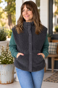 A woman wearing a dark gray sherpa fleece button-front vest over a teal long-sleeve shirt and blue jeans, standing on a porch with plants and furniture in the background.