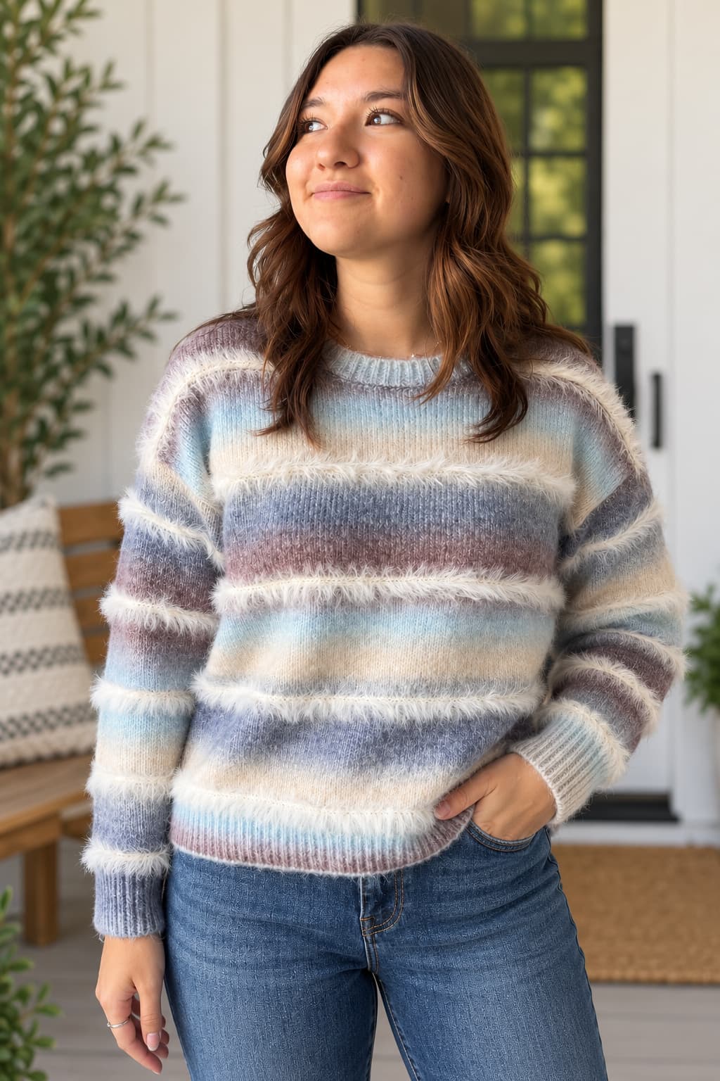 A woman wearing a fuzzy, horizontally striped knit sweater in pastel shades of blue, purple, beige, and white, paired with blue jeans, standing on a porch.