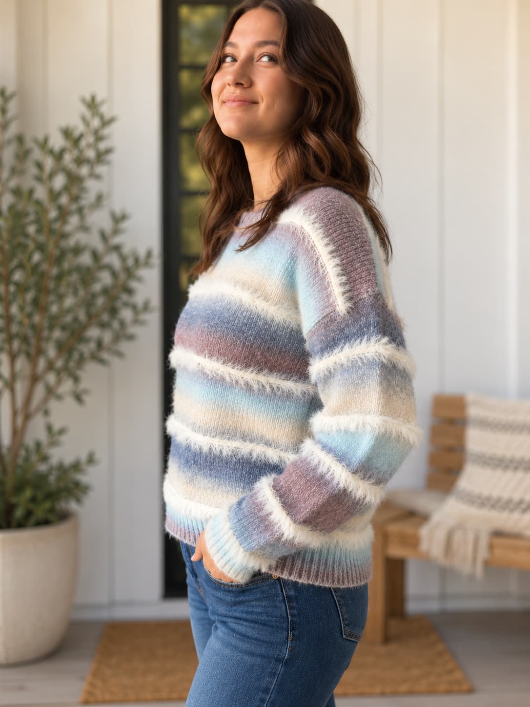 A woman wearing a fuzzy, long-sleeve knit sweater with horizontal pastel stripes in blue, purple, pink, and cream, paired with blue jeans, standing in a bright, modern porch setting.