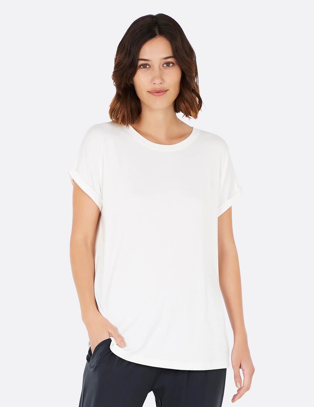 A woman with shoulder-length brown hair wearing a white short-sleeve crewneck t-shirt and dark pants, standing against a light gray studio background.