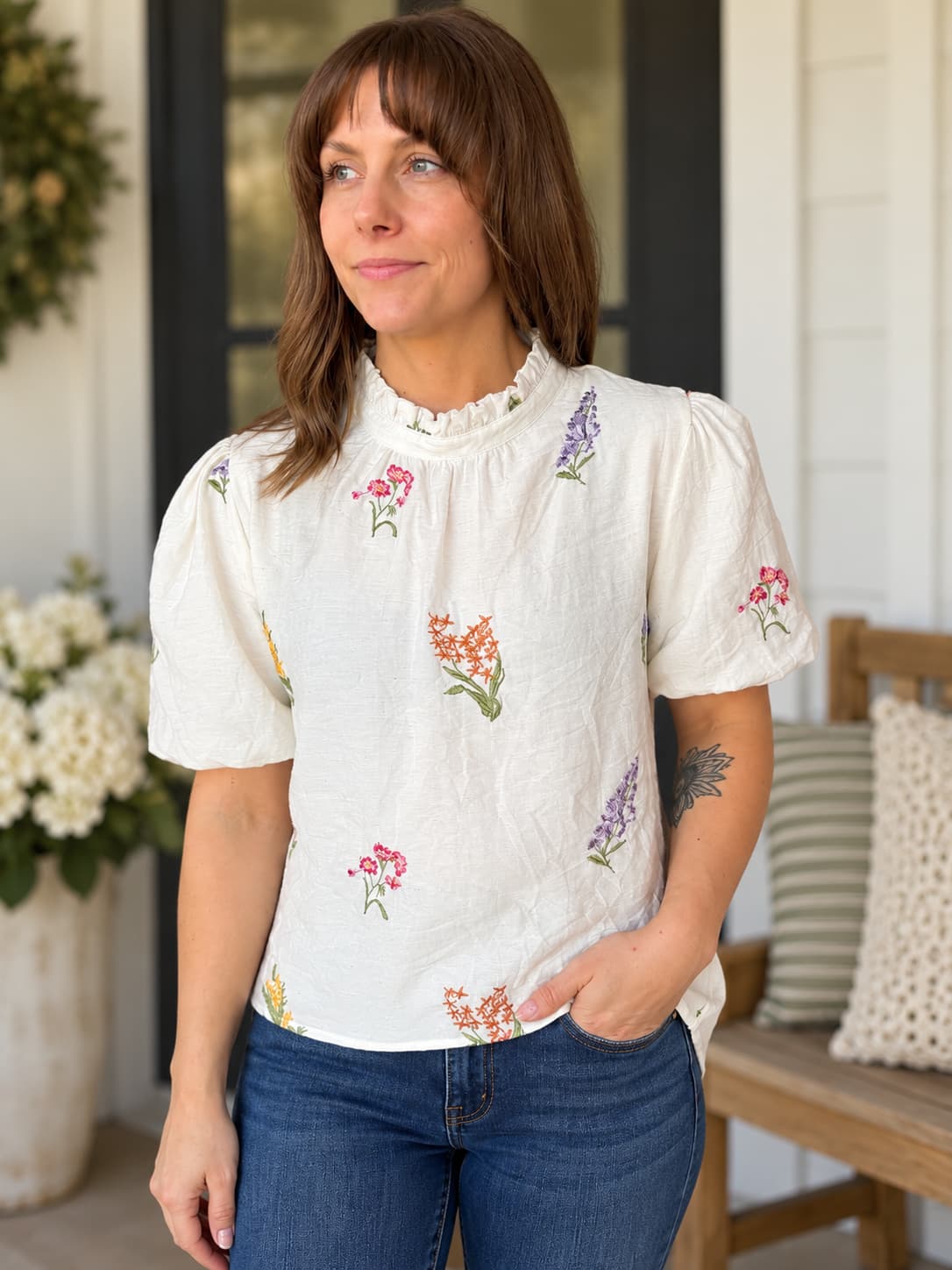 A woman wearing a short-sleeve white blouse with colorful floral embroidery and blue jeans, standing on a porch.