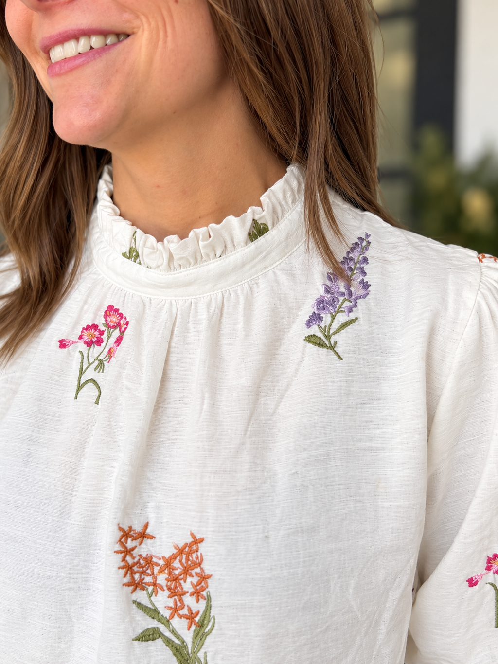 Close-up of a woman wearing a white linen blouse with colorful floral embroidery, featuring a ruffled mock neck and puff sleeves.