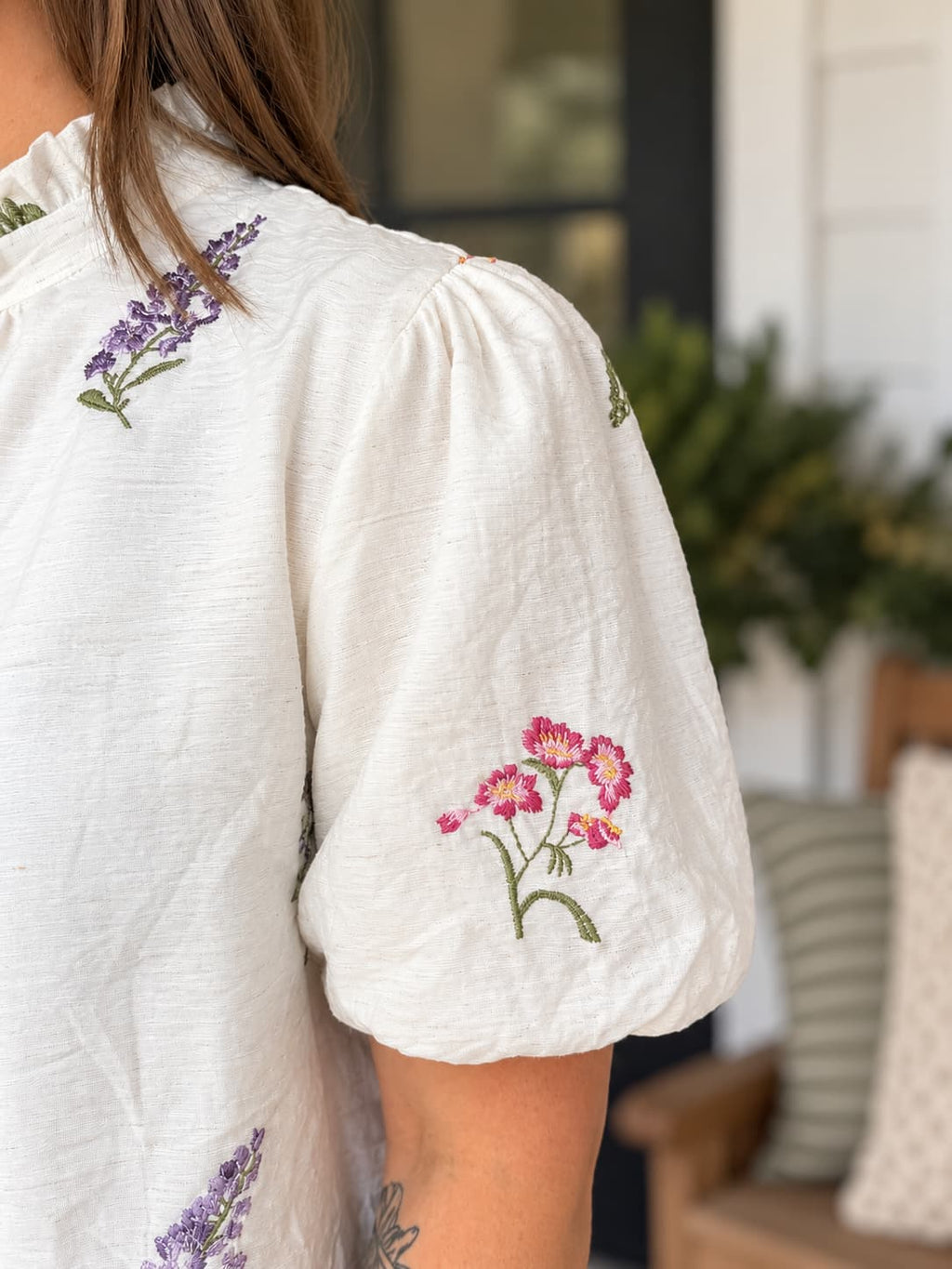 Close-up of a person wearing a white short-sleeve blouse with colorful floral embroidery on the shoulder and sleeve, shown in an outdoor patio setting.