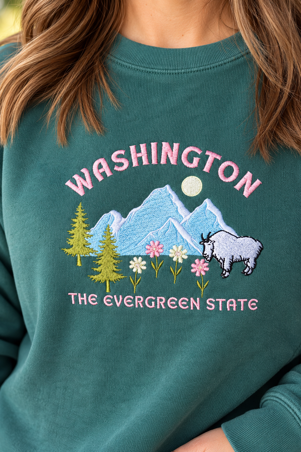 A close-up of a person wearing a teal crewneck sweatshirt with a colorful embroidered graphic of mountains, trees, flowers, and a goat, with the text 'WASHINGTON THE EVERGREEN STATE'.
