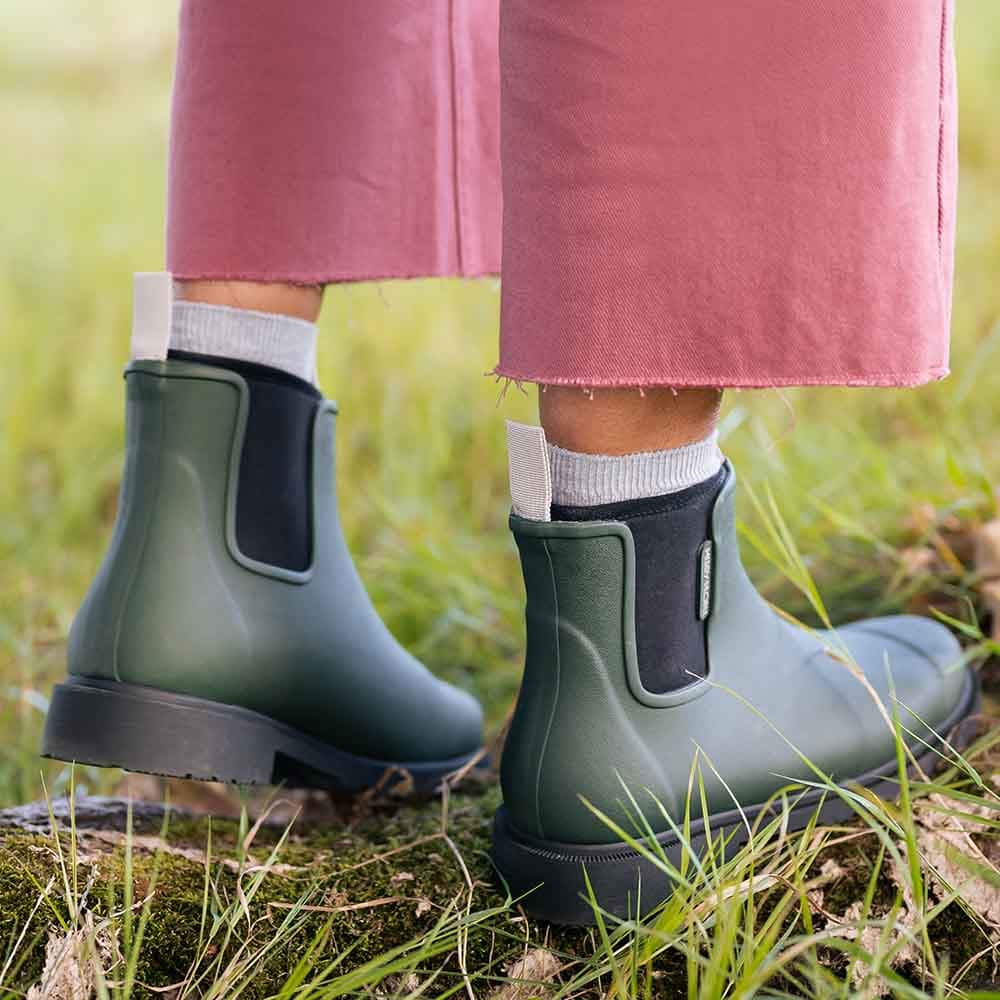 A person wearing dark green Chelsea rain boots with black elastic side panels, paired with pink trousers and white socks, standing outdoors on grass and moss.