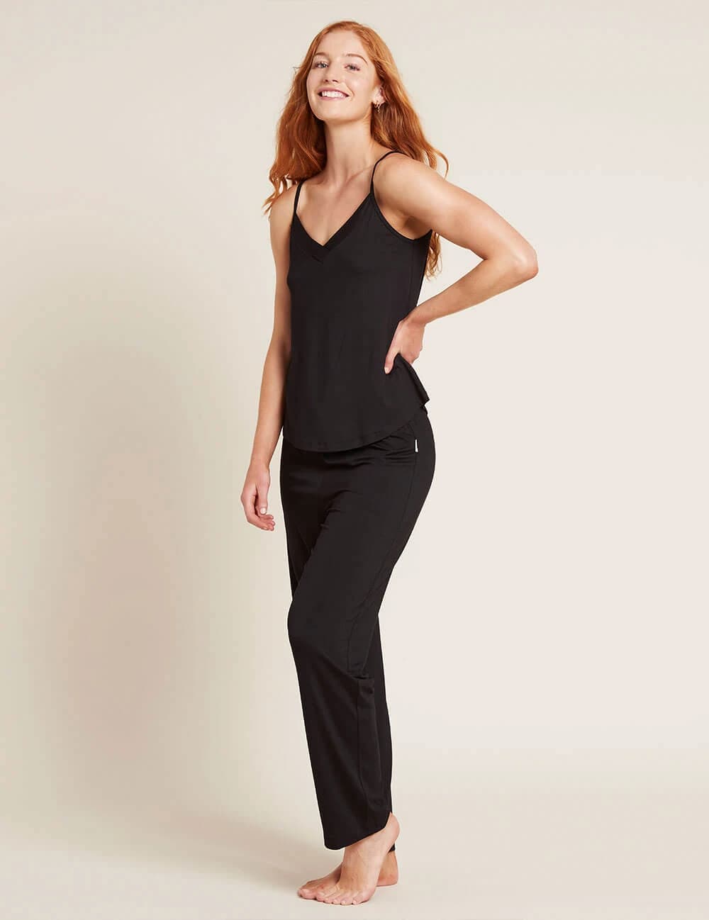 A woman modeling a black camisole top and matching black pants against a plain light beige studio background.