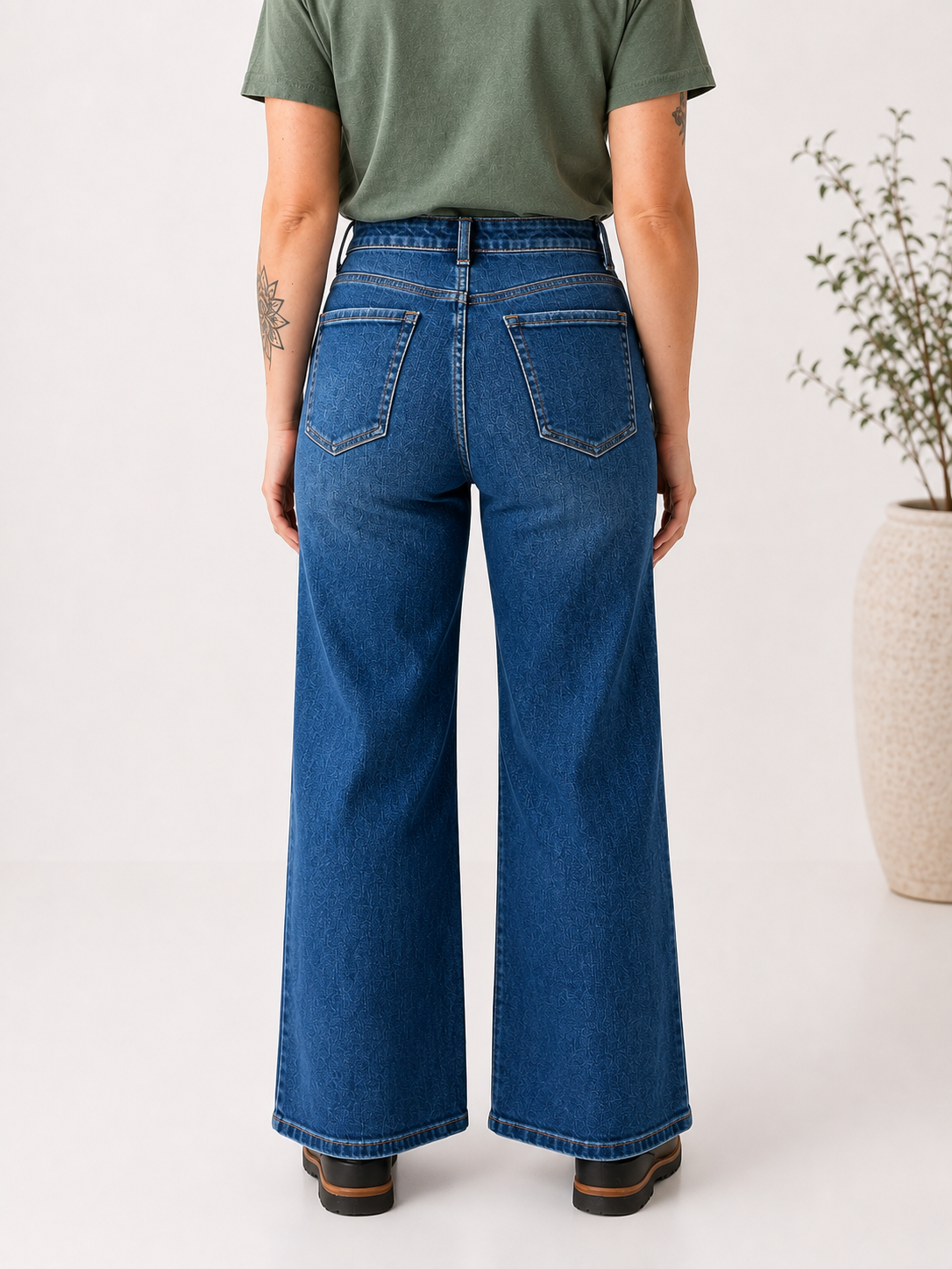 A person shown from the back wearing blue wide-leg denim jeans, an olive green t-shirt, and brown shoes, standing in a studio setting with a potted plant in the background.