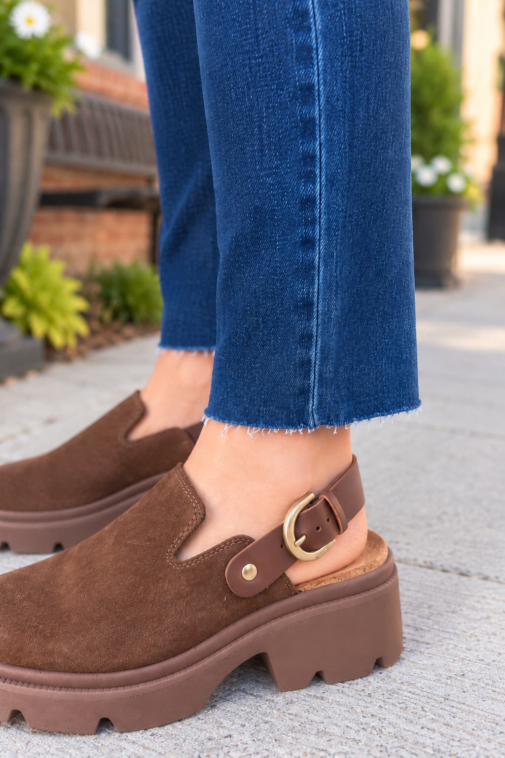 A close-up of a person wearing brown suede clog-style mules with platform soles and ankle straps, paired with blue jeans on a sidewalk.