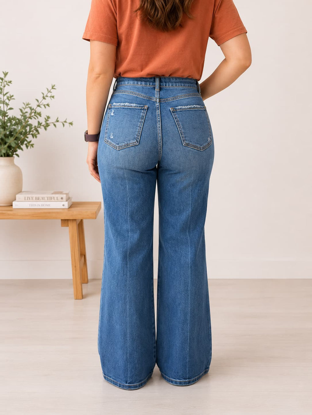 A person shown from the back wearing blue wide-leg jeans and an orange short-sleeve top in a bright indoor room with a small wooden side table and plant.