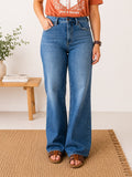 A person wearing high-waisted, wide-leg blue denim jeans with a faded wash, paired with a rust-orange graphic t-shirt and brown leather slide sandals, standing on a woven jute rug.