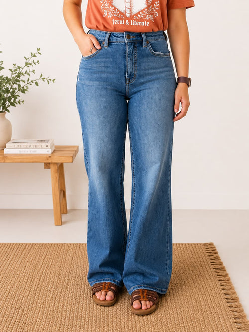A person wearing high-waisted, wide-leg blue denim jeans with a faded wash, paired with a rust-orange graphic t-shirt and brown leather slide sandals, standing on a woven jute rug.
