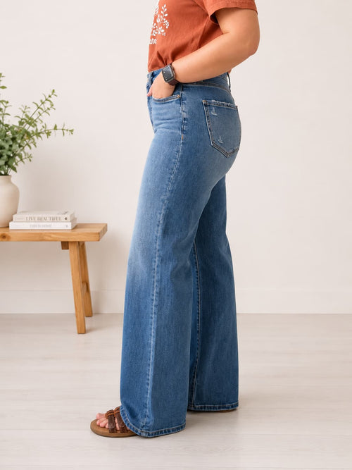 A person wearing blue wide-leg jeans, an orange t-shirt, and brown sandals, standing in a minimalist room with a small wooden side table and a plant.
