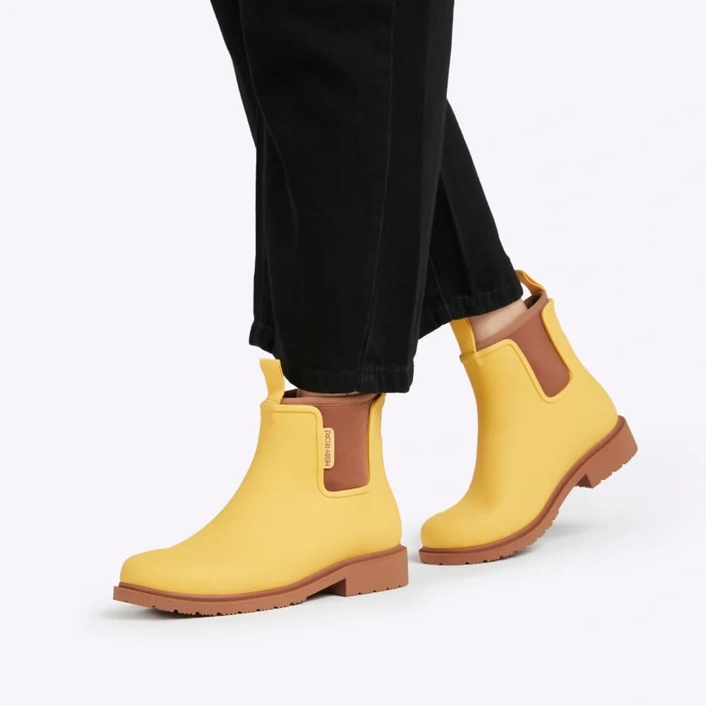 A person wearing bright yellow Chelsea-style rain boots with brown elastic side panels and brown soles, paired with black pants, on a clean white studio background.