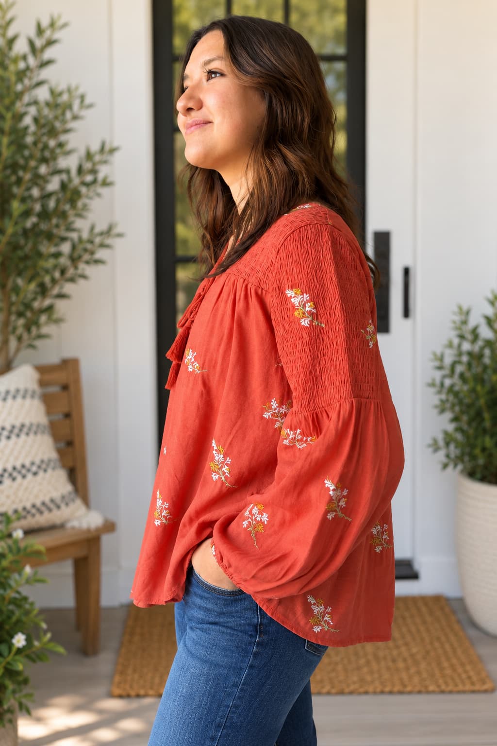 A woman wearing a rust-orange off-the-shoulder blouse with white floral embroidery and blue jeans, standing in a bright doorway with plants and a wooden chair in the background.