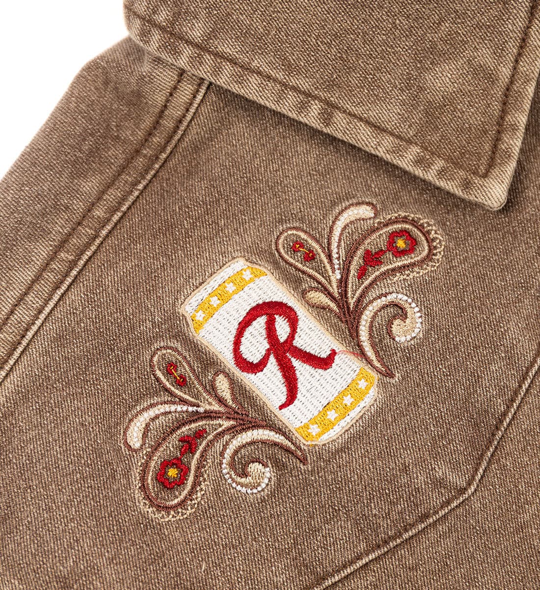 Close-up of a brown denim jacket pocket showing an embroidered monogram patch with a red letter 'R' on a white and yellow background, surrounded by brown and red paisley embroidery.