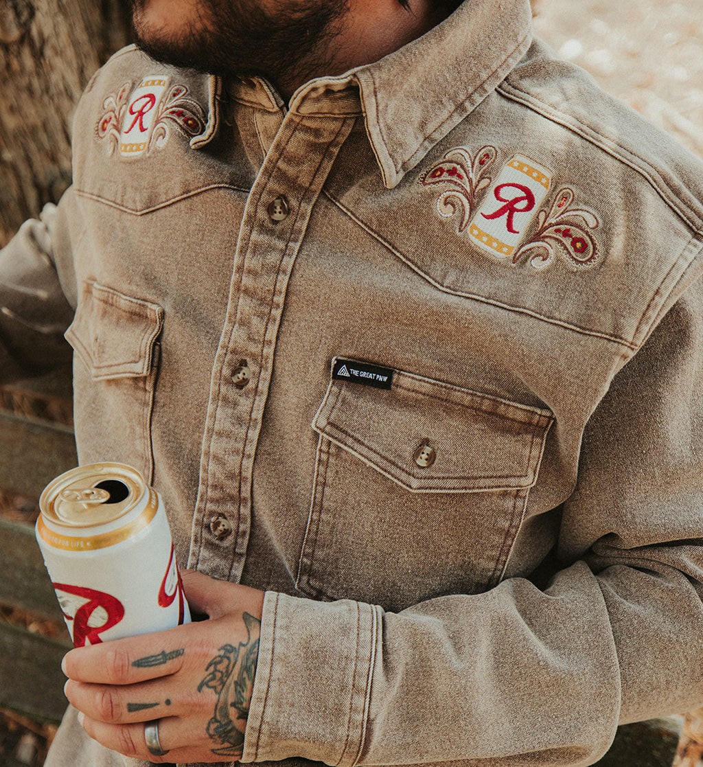A close-up of a man wearing a tan western-style denim shirt jacket with embroidered patches on the chest, holding a white and gold beer can.