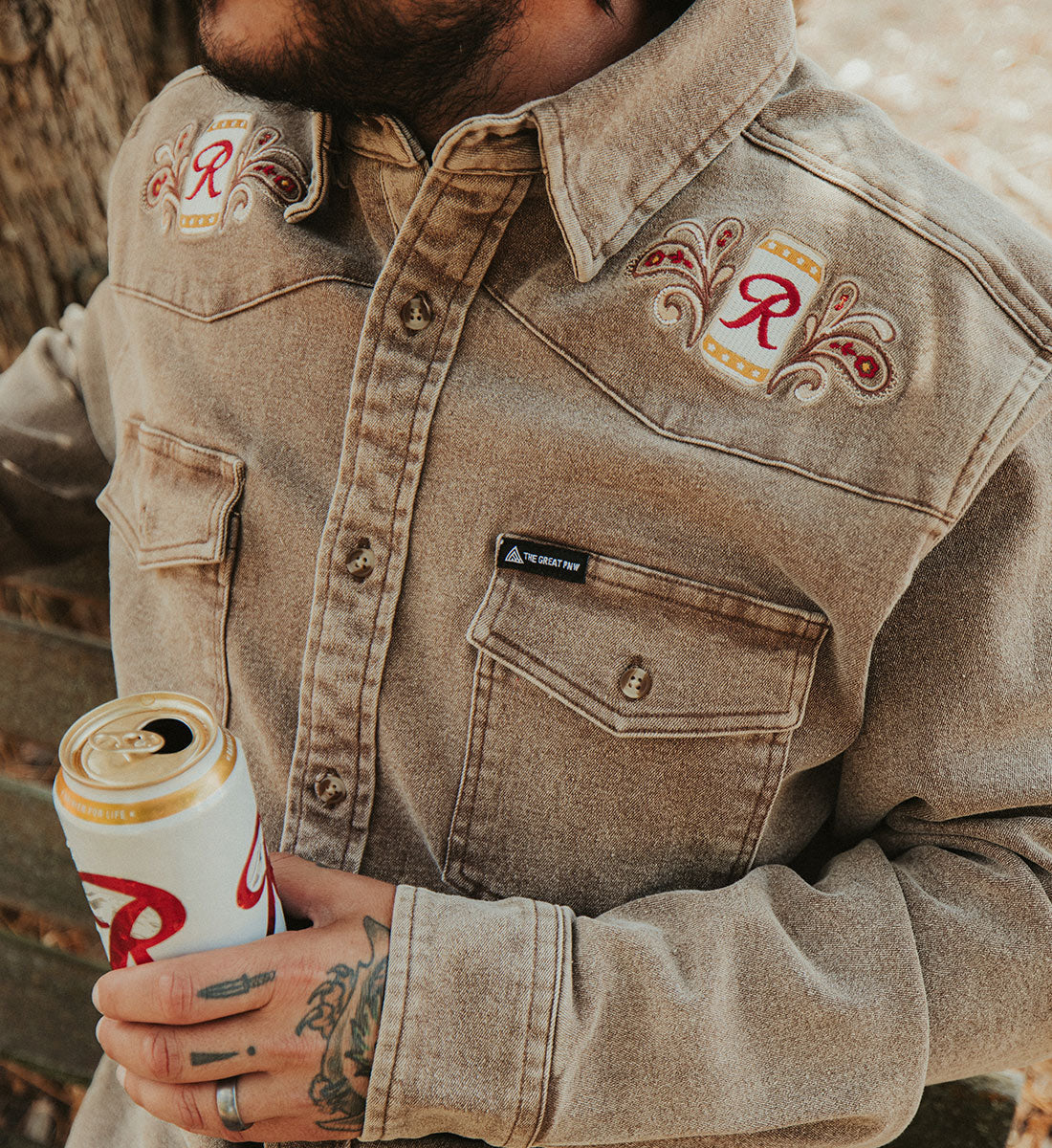 A close-up of a man wearing a tan western-style denim shirt jacket with embroidered patches on the chest, holding a white and gold beer can.