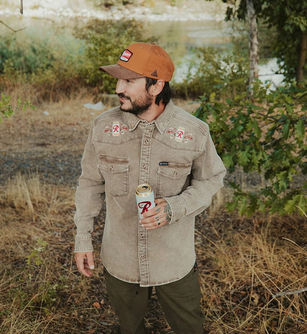 A man wearing a beige western-style button-up shirt with embroidered details on the chest, an orange-brown cap, and olive pants, holding a can of beer outdoors near a lake.