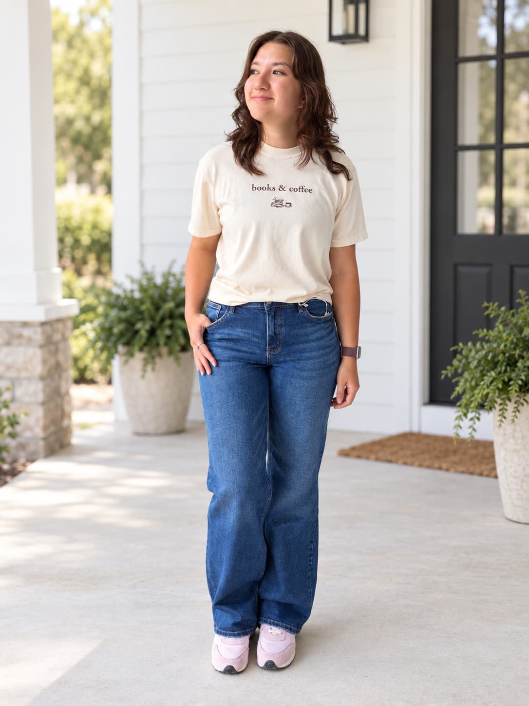 A woman stands on a front porch wearing a cream graphic t-shirt with 'books & coffee' printed on the chest, blue jeans, and pink sneakers.
