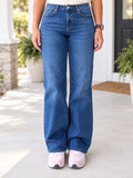 A person wearing a pair of medium-wash blue straight-leg jeans, standing on a concrete porch with potted plants and a white door in the background.