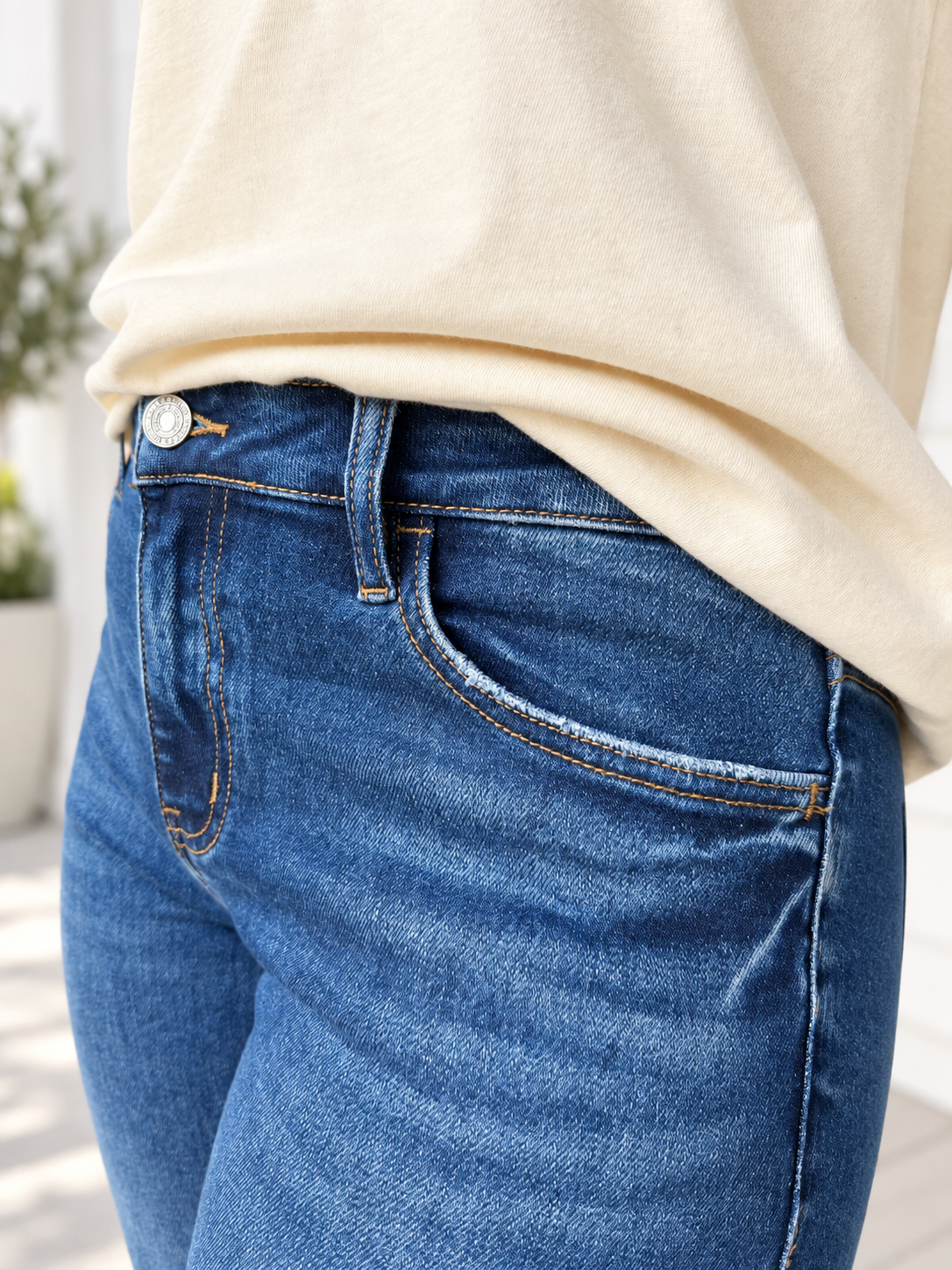 Close-up of a person wearing medium-wash blue denim jeans with a cream-colored top, showing the front pocket and waistband details.