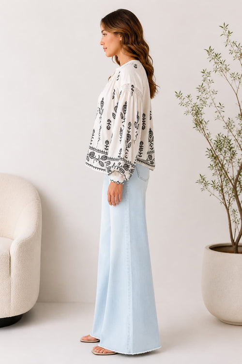 Woman wearing a white blouse with black embroidery patterns and light blue pants in a minimalistic room.