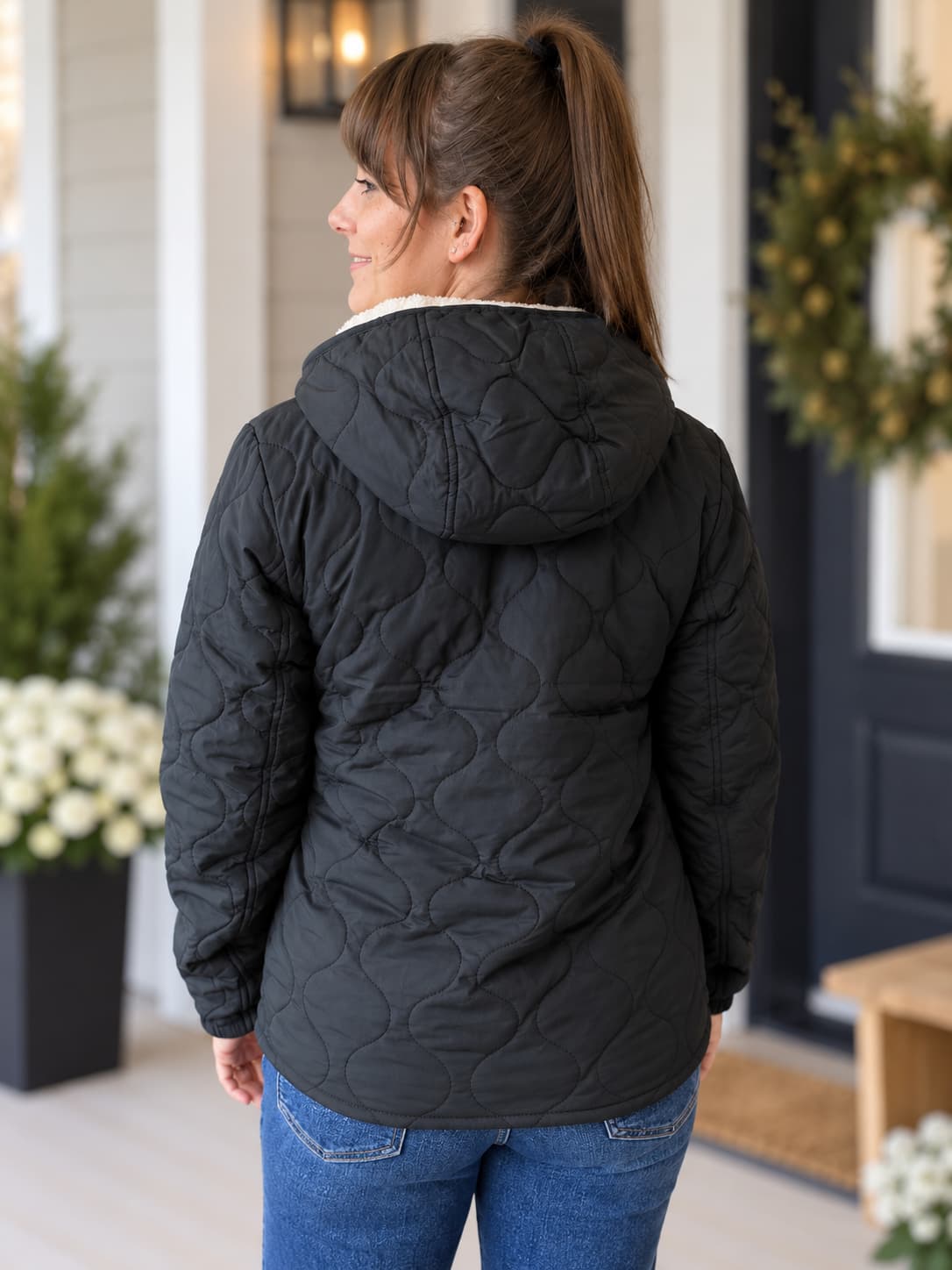 A woman viewed from the back wearing a black quilted hooded jacket with a white sherpa lining, paired with blue jeans, standing on a front porch decorated with greenery and flowers.
