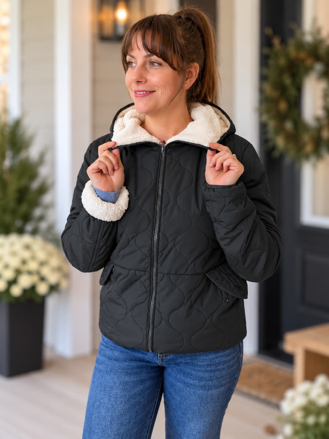 A woman wearing a black quilted puffer jacket with a cream sherpa-lined hood and cuffs, paired with blue jeans, standing on a front porch.