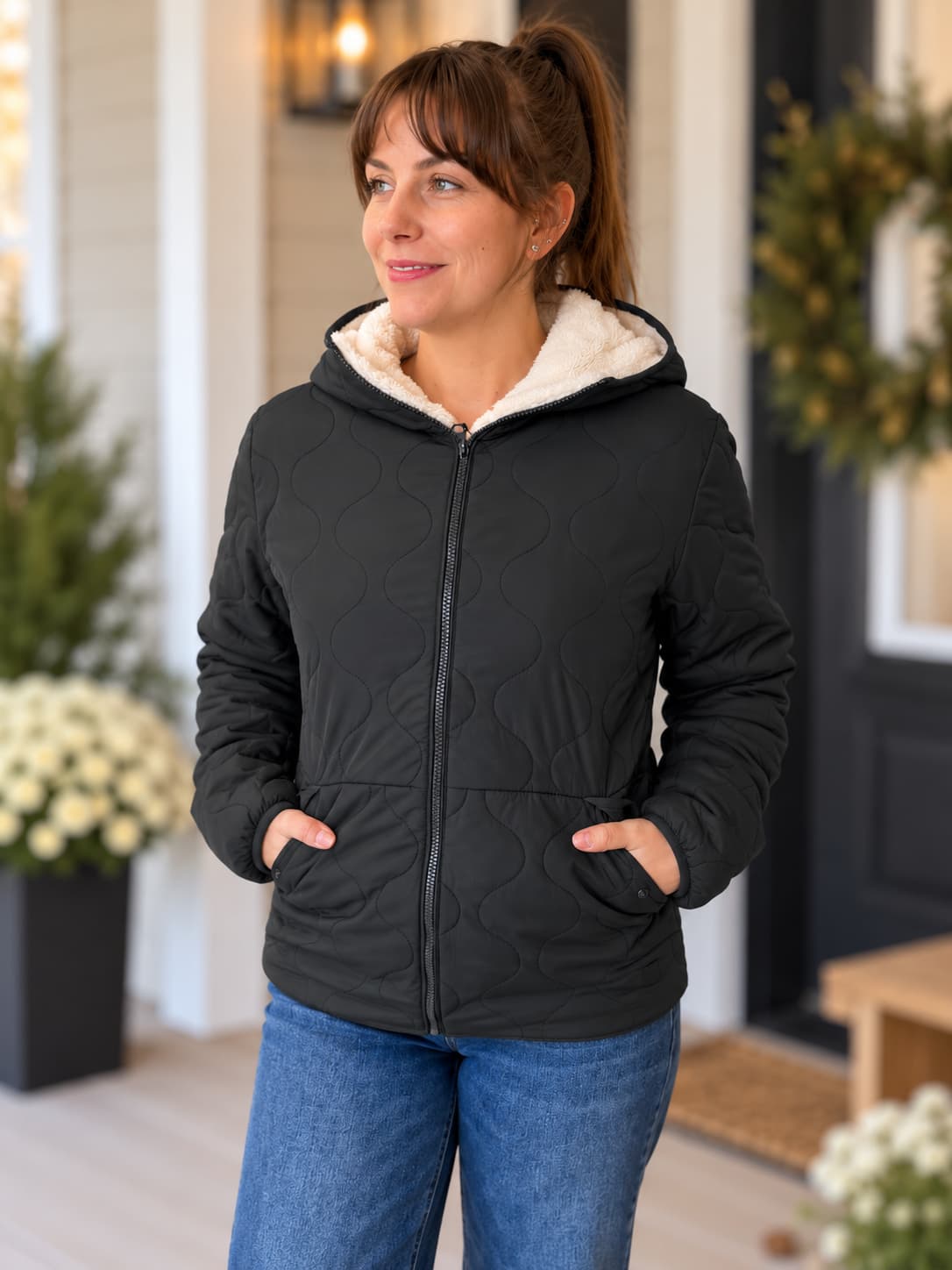 A woman wearing a black quilted zip-up hooded jacket with a cream sherpa-lined hood and blue jeans, standing on a front porch.