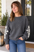 A woman wearing a dark gray crewneck sweater with white floral embroidery on the sleeves, paired with blue jeans, standing on a front porch.