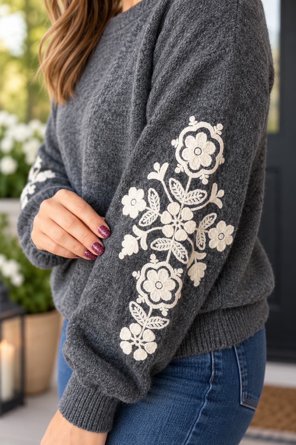A woman wearing a dark gray knit sweater with large cream floral embroidery on the sleeve, paired with blue jeans in an outdoor setting.