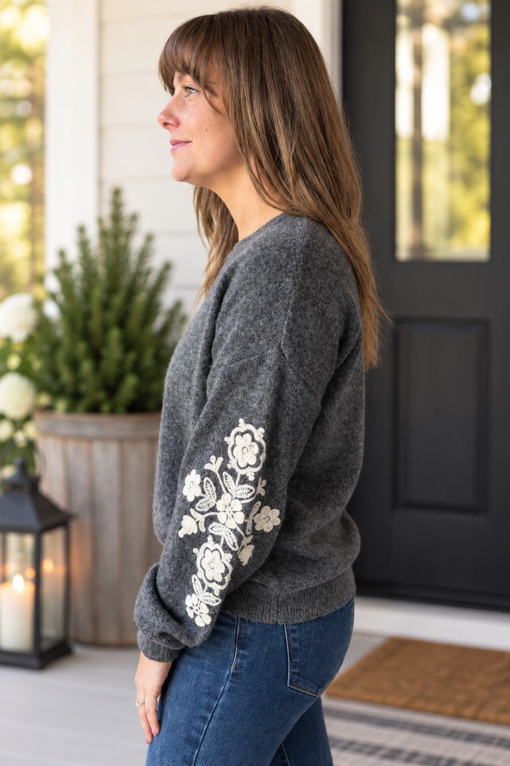 A woman wearing a dark gray knit sweater with white floral embroidery on the sleeve, standing in profile on a front porch.