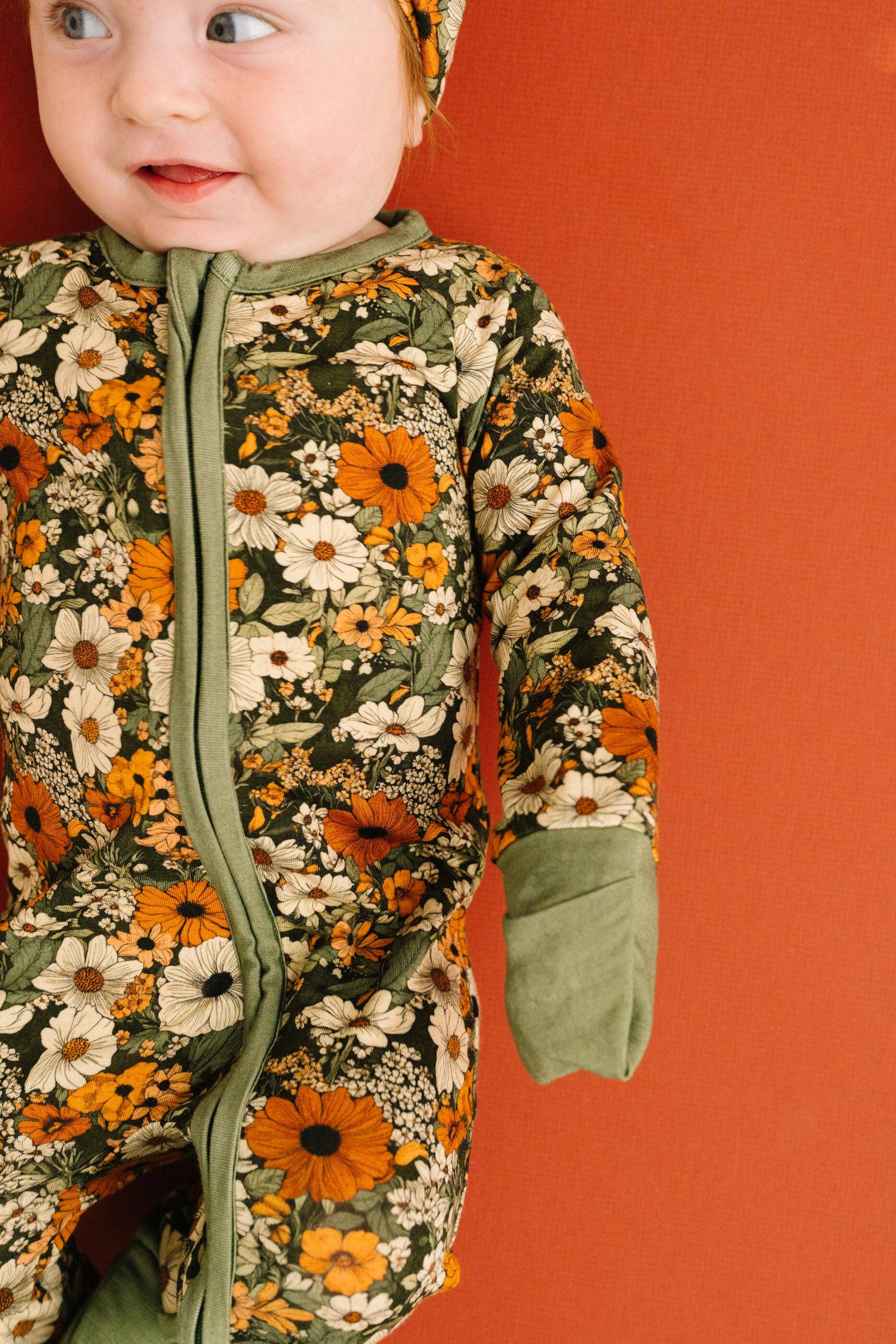 A baby wearing a floral print zip-up sleeper with sage green trim, standing against a solid terracotta orange background.