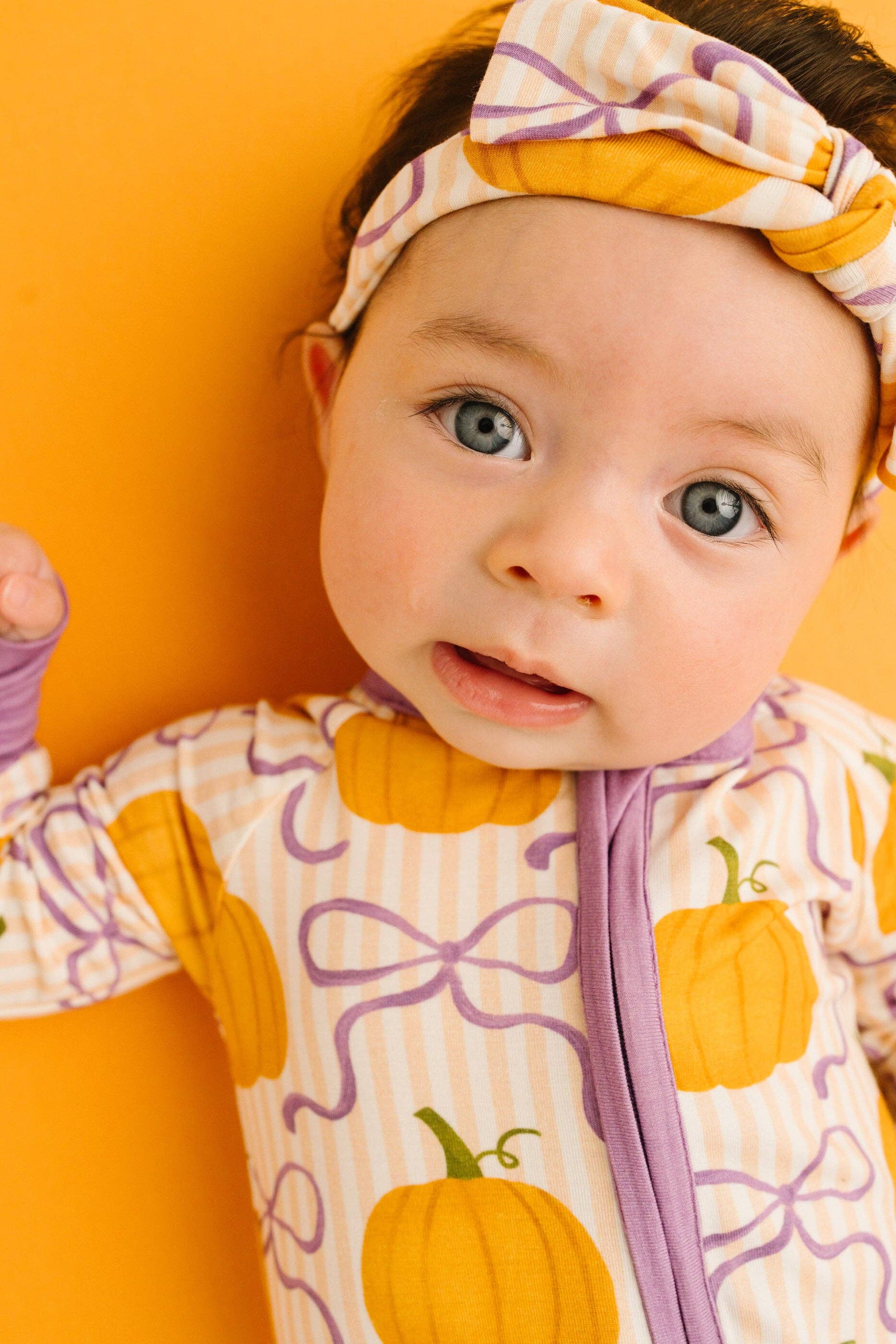 A baby wearing a cozy Halloween-themed footless onesie and matching headband, featuring a pumpkin and bow pattern in orange, yellow, and purple on a cream background, set against a solid orange backdrop.