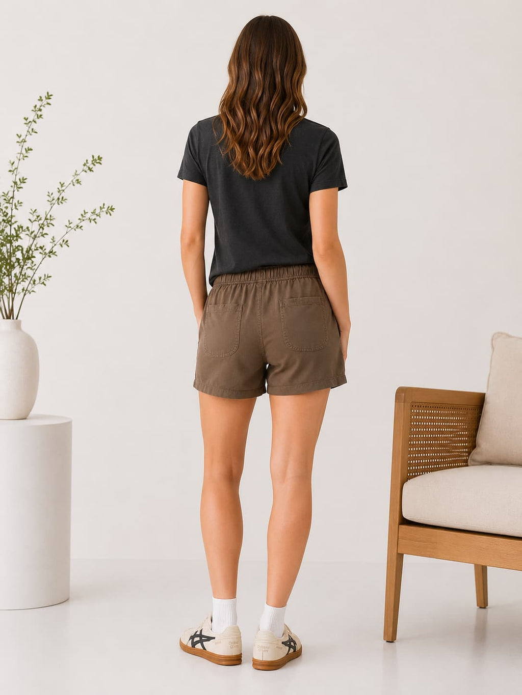 Woman wearing a black t-shirt and brown tencel shorts standing in a minimalistic room with a plant and chair.
