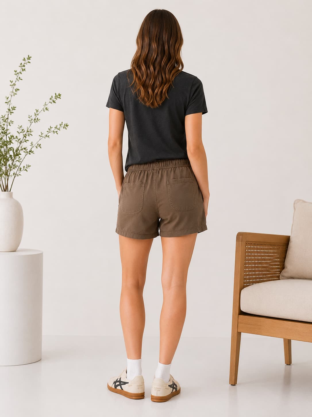Woman wearing a black t-shirt and brown tencel shorts standing in a minimalistic room with a plant and chair.