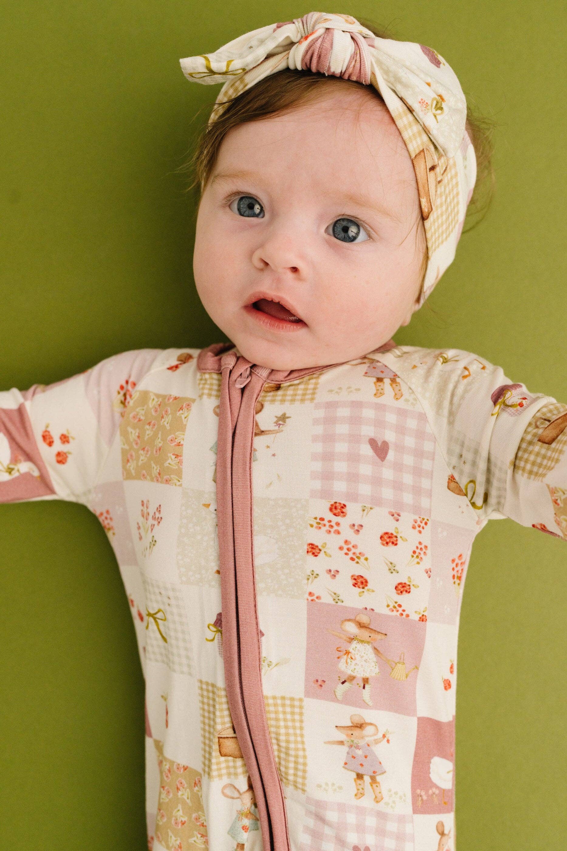 Baby wearing a patchwork bamboo sleeper with a floral headband against a green background