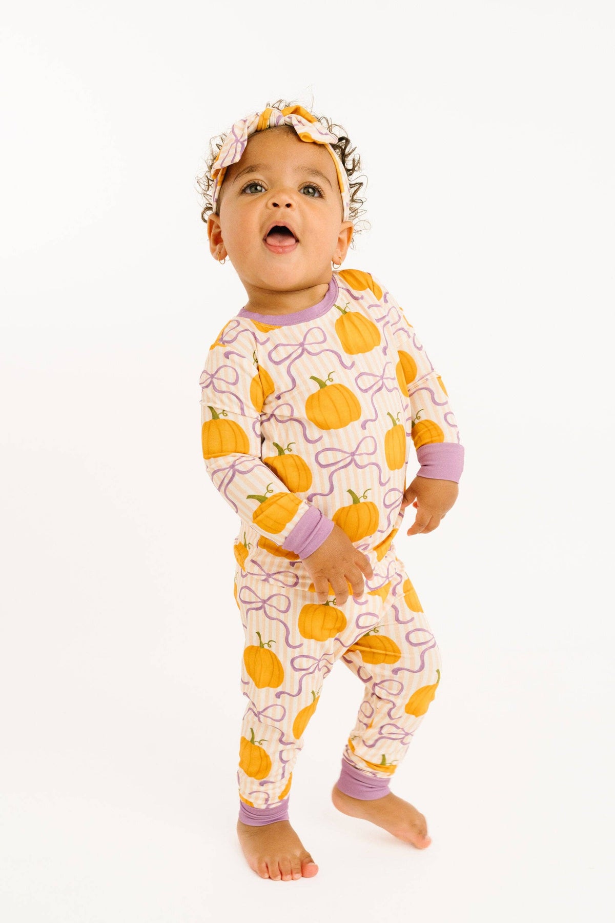 A baby standing barefoot on a white studio background, wearing a long-sleeve pajama set with an orange pumpkin and purple bow pattern, along with a matching headband.